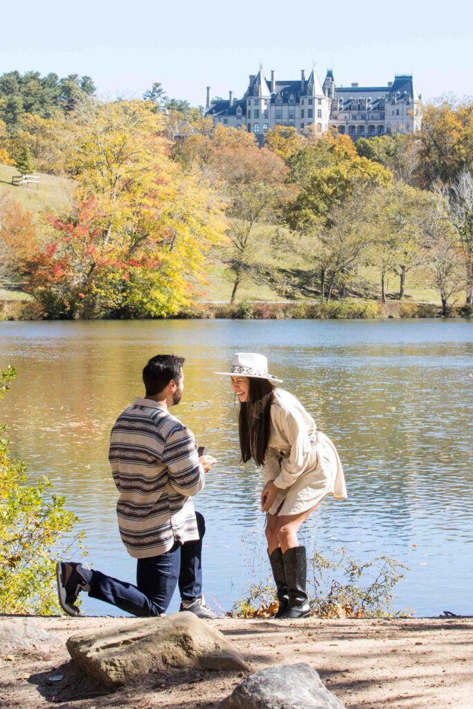 woman leans forward in excitement as her fiance proposes to her in front of a lake at the biltmore estate