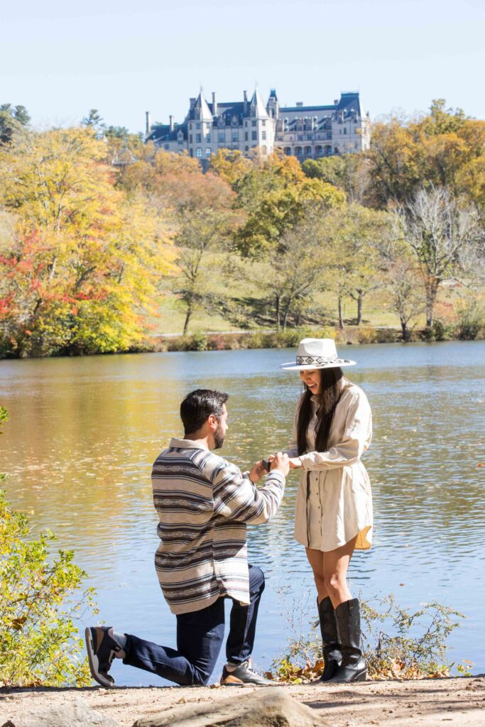 man proposes on one knee to his girlfriend at the lagoon with the biltmore estate in the background