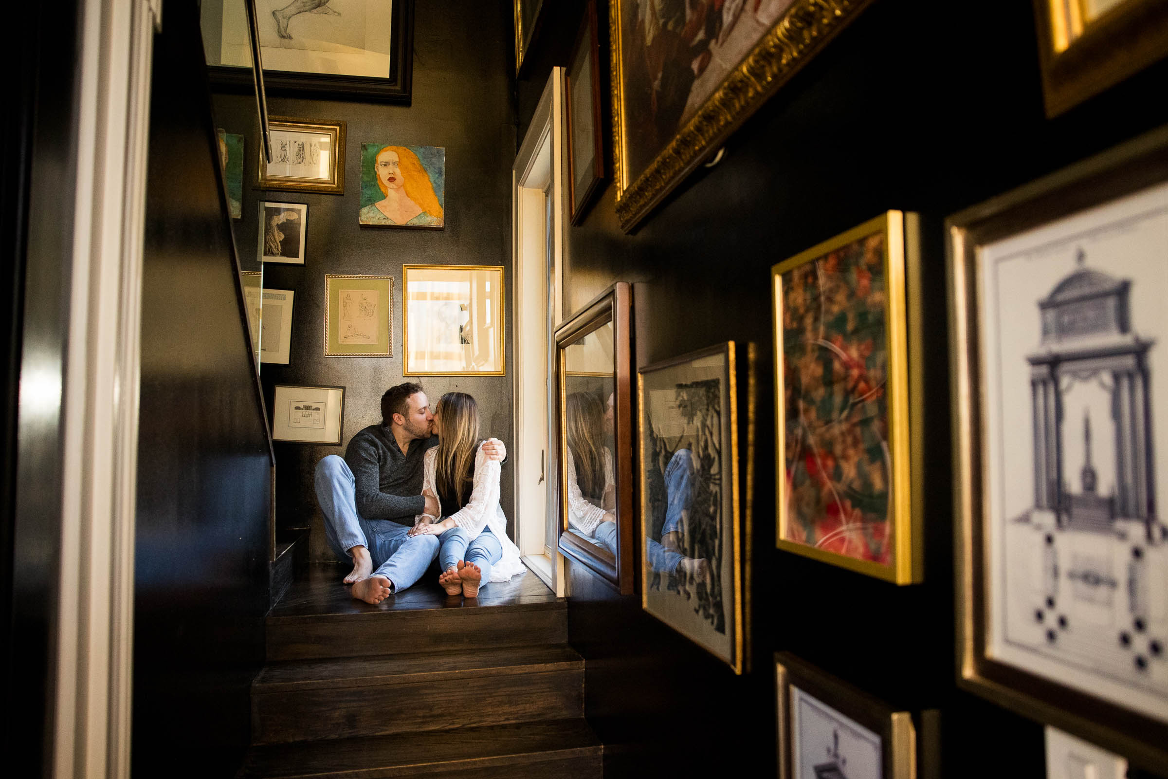 couple kisses while sitting on the stairs in a hallway filled with framed artwork at the Redstone Mansion in Burnsville during an Airbnb session the evening before their NC elopement
