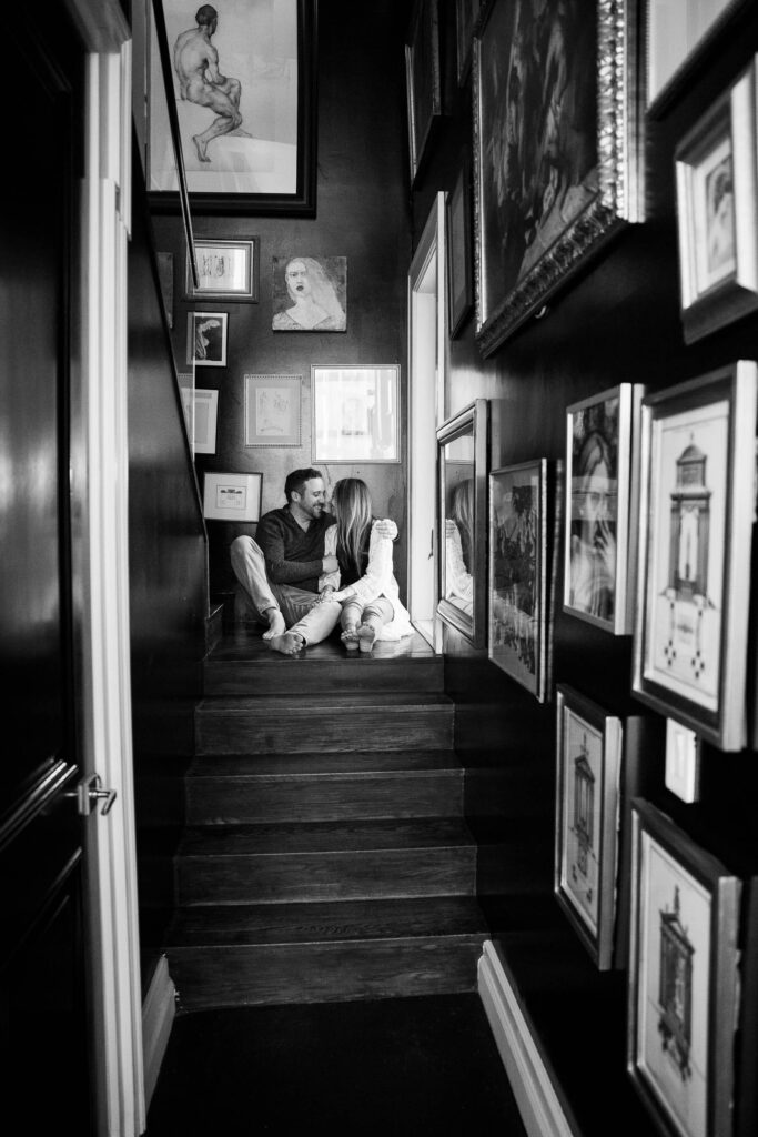 couple kisses while sitting in a hallway lined with framed art in a black and white photo 