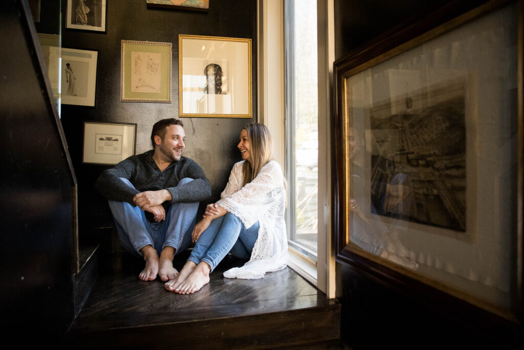 couple flirts while sitting barefoot on the floor in the hallway of their airbnb, surrounded by framed artwork