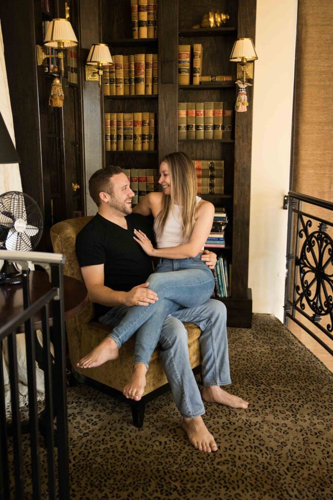 women sits on her fiances lap in a library at their funky airbnb in north carolina