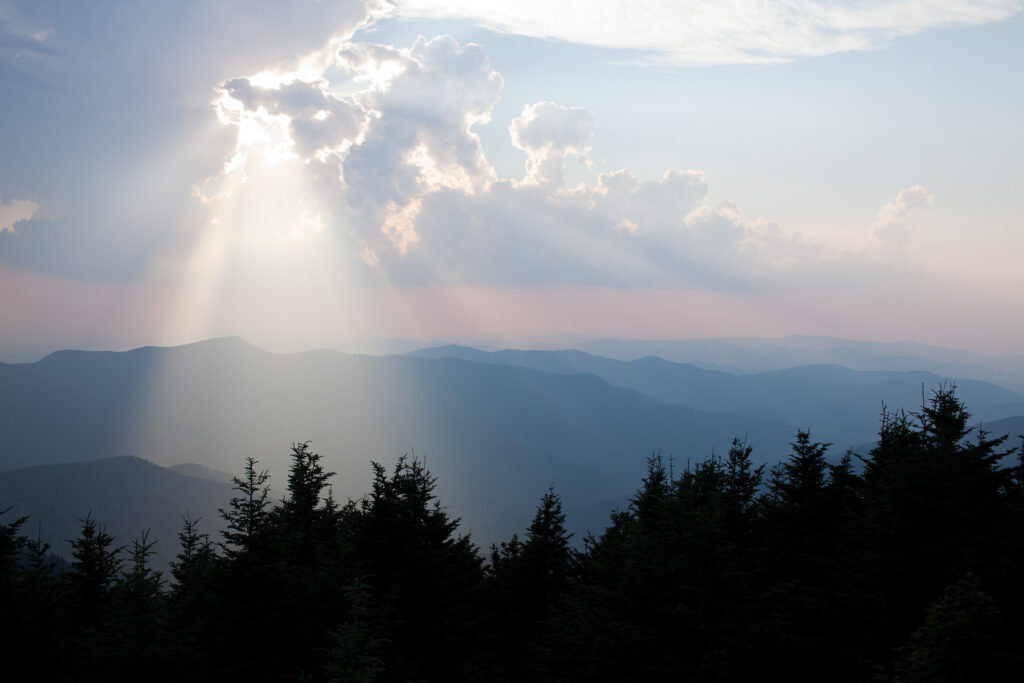 beams of sunlight shine down through storm clouds onto evergreen trees, silouetted against the blue ridge mountains and sunset sky at mount mitchell in north carolina along the blue ridge parkway
