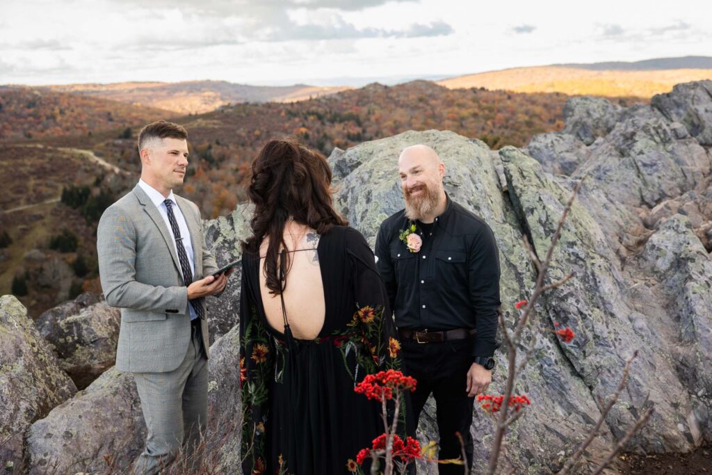 groom wearing all black smiles at the bride as the officiant looks on during vows on a mountain at grayson highlands in va