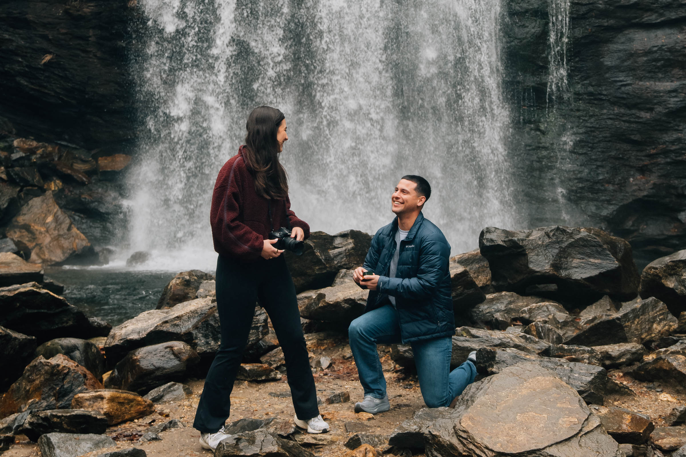 Surprise marriage proposal at Looking Glass Falls in Pisgah National Forest near Brevard, North Carolina, with the groom kneeling on river rocks beneath the waterfall.