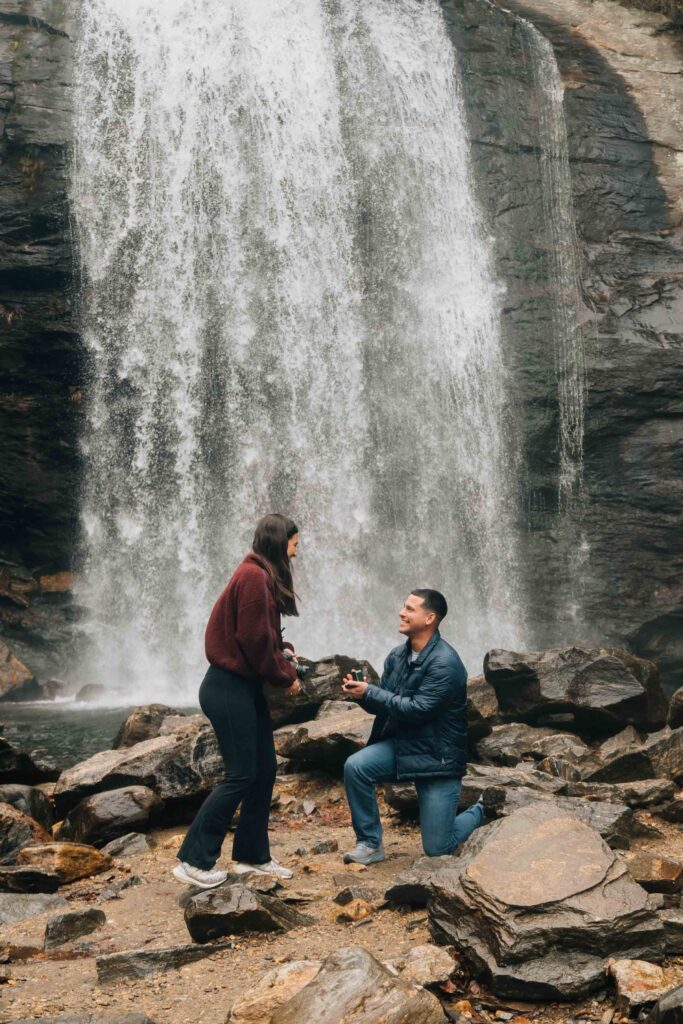Surprise marriage proposal at Looking Glass Falls in Pisgah National Forest near Brevard, North Carolina, with the groom kneeling on river rocks beneath the waterfall.