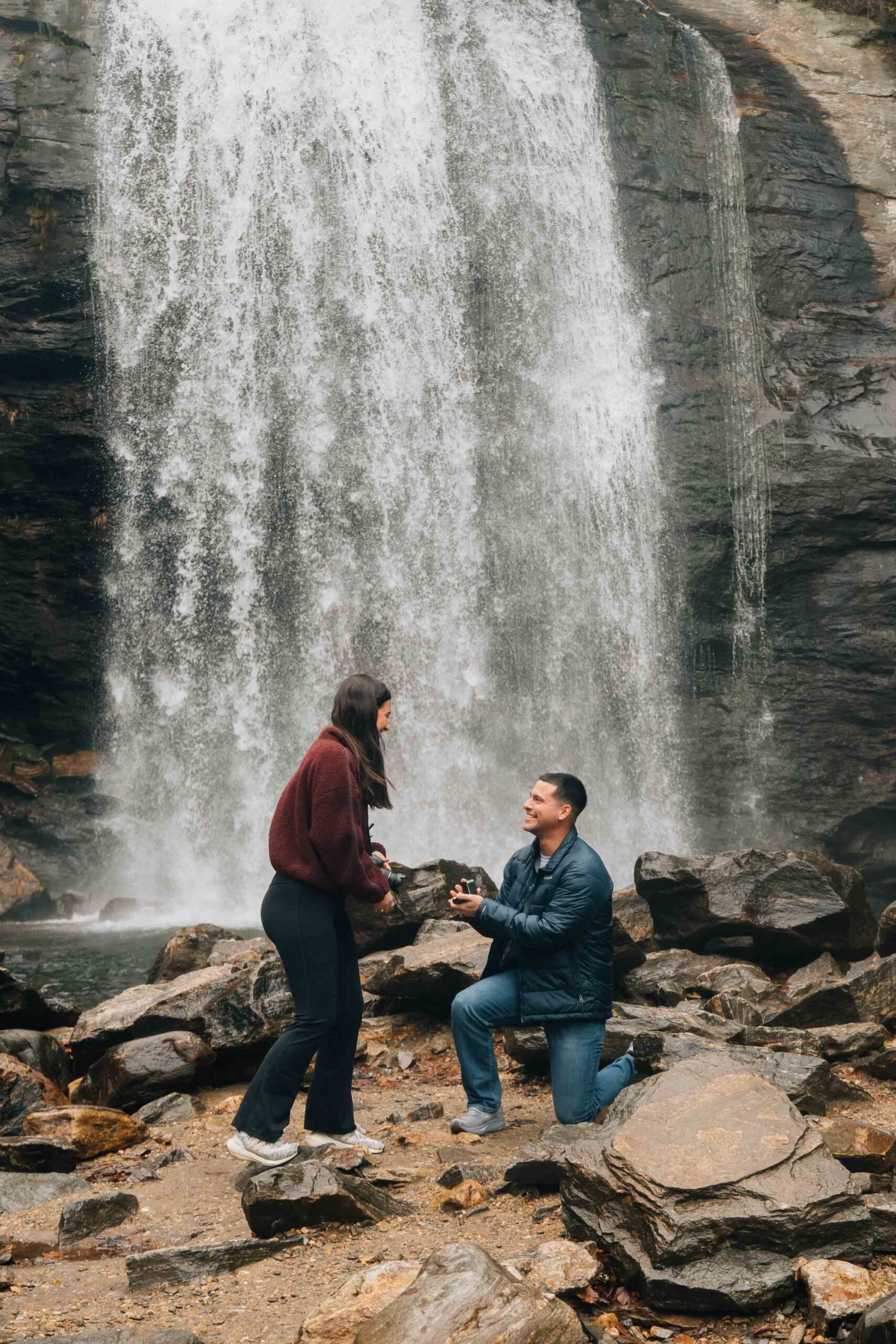 Surprise marriage proposal at Looking Glass Falls in Pisgah National Forest near Brevard, North Carolina, with the groom kneeling on river rocks beneath the waterfall.
