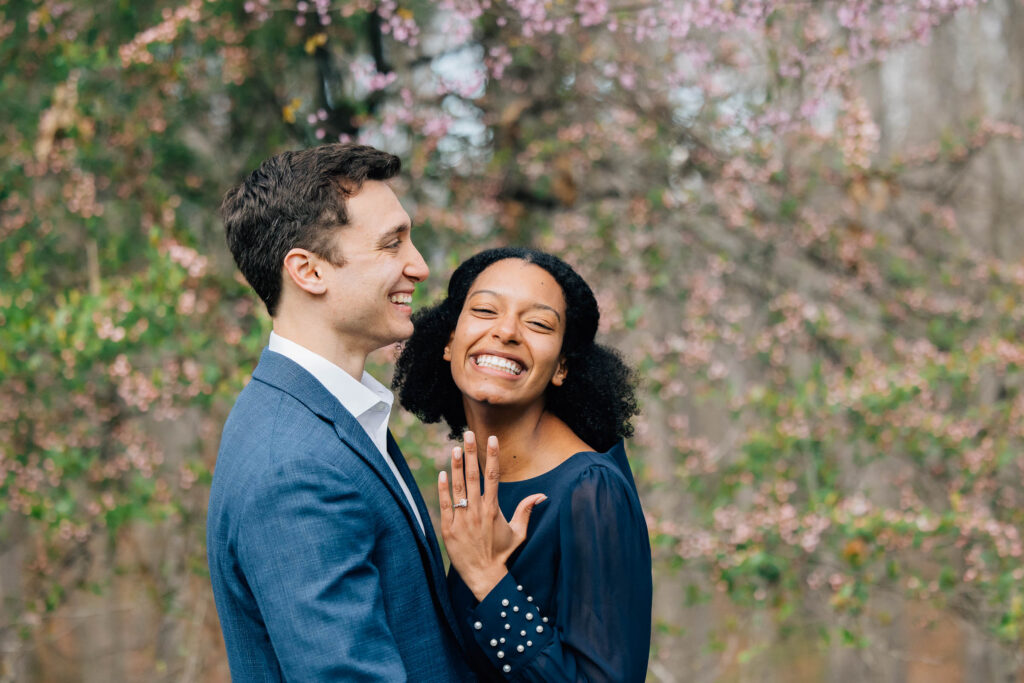 woman holds up her hand with her new engagement ring in asheville at the unc arboretum after their proposal