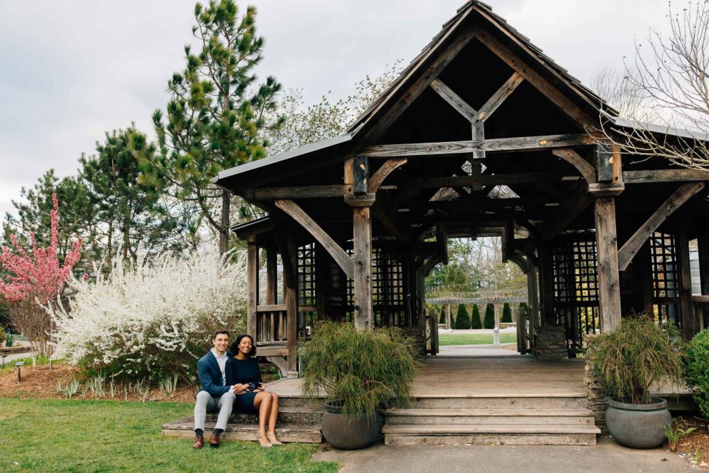 couple sits under a timber frame pergola at the unc arboretum in asheville during their engagement session after their proposal