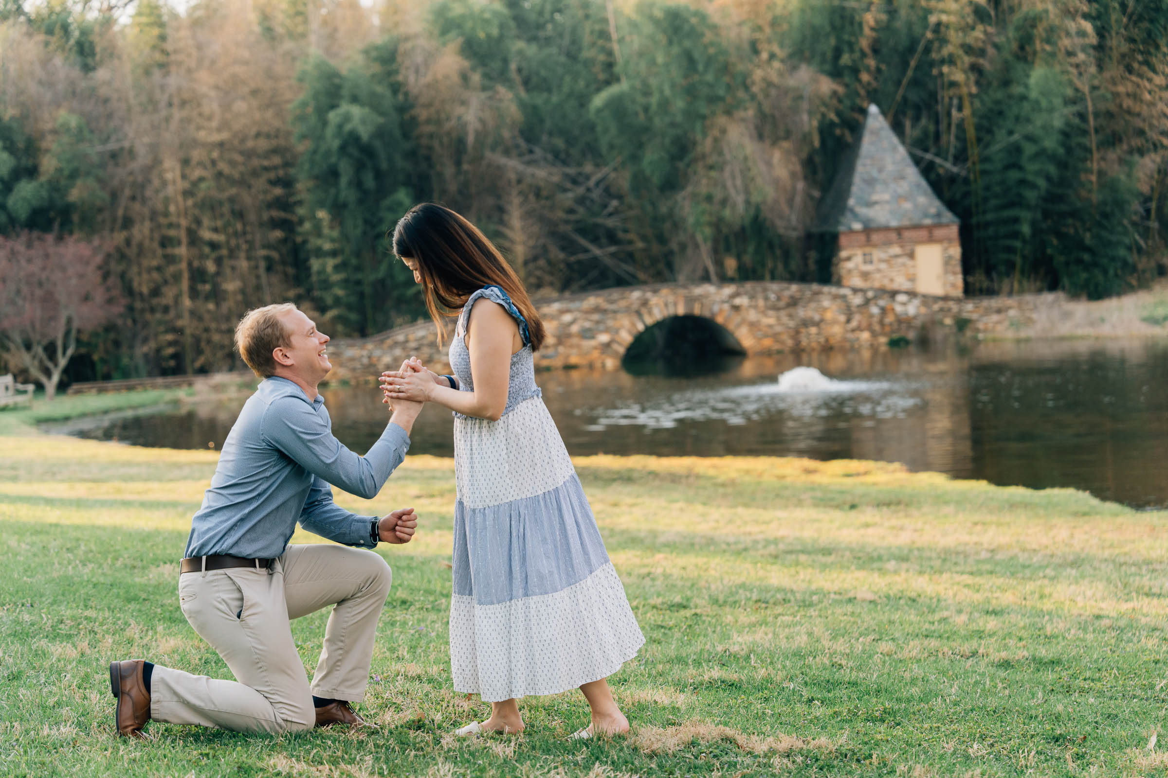 man on one knee, giving ring to his partner during a surprise proposal at the graylyn estate in winston salem, nc with the pond and bridge in the background