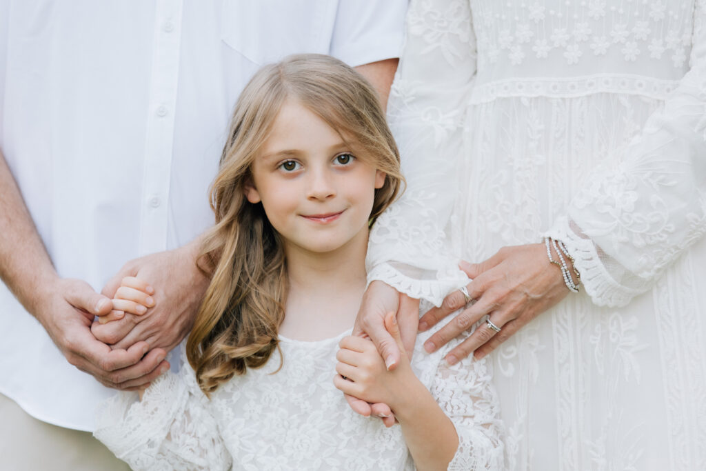 little girl in white tenderly holds hands with her parents and smiles at the camera during a family session at hurley park in salisbury north carolina