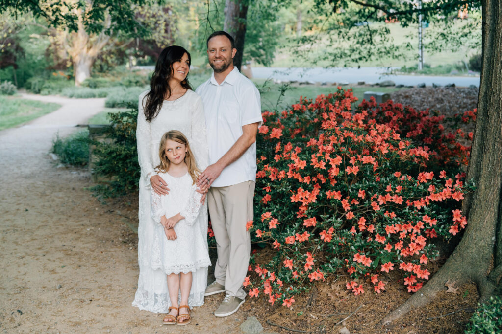 girl makes silly face while standing with her parents in front of azaleas in the springtime in hurley park in salisbury nc
