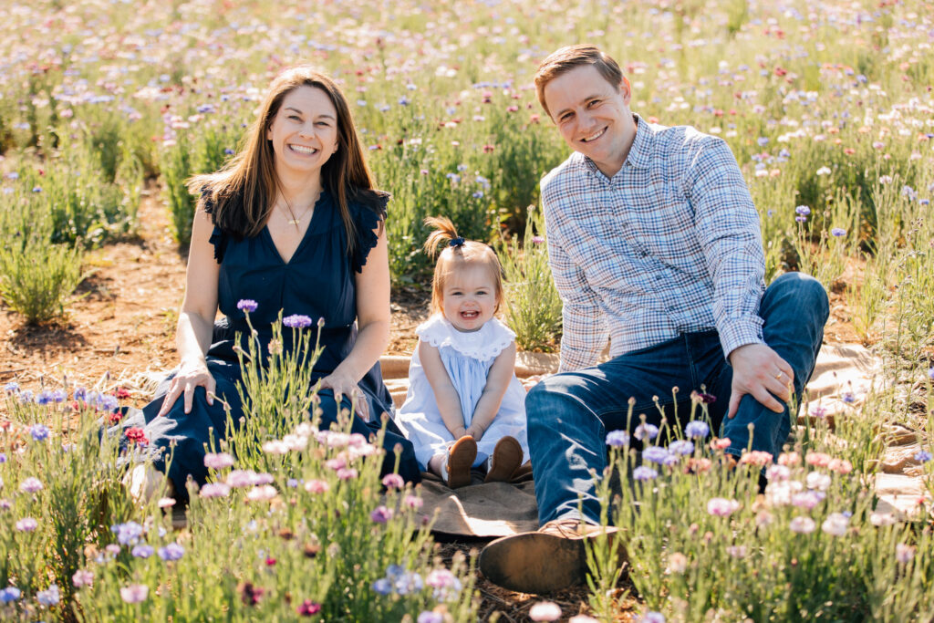 cute toddler with a tiny pony tail sits between her parents in a field of wildflowers in north carolina