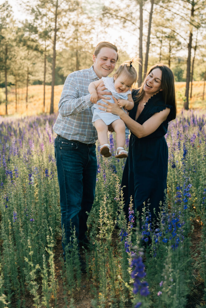 parents hug and swing their toddler daughter in a field of wildflowers at dogwood farms in belews creek, north carolina