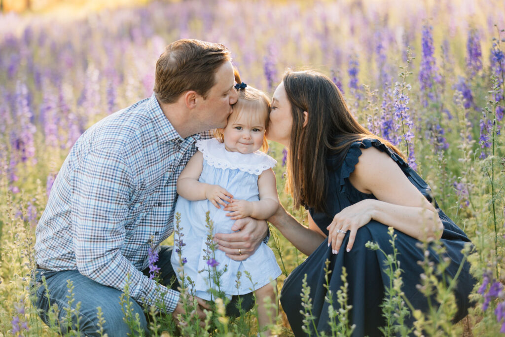 mom and dad kiss adorable toddler while sitting in a field of purple flowers at dogwood farms in belews creek, nc