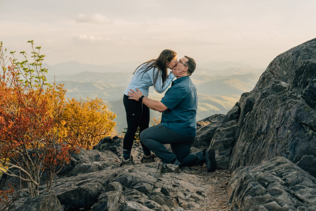 woman kisses her boyfriend as he proposes on on knee with a mountain backdrop at grayson highlands state park in va