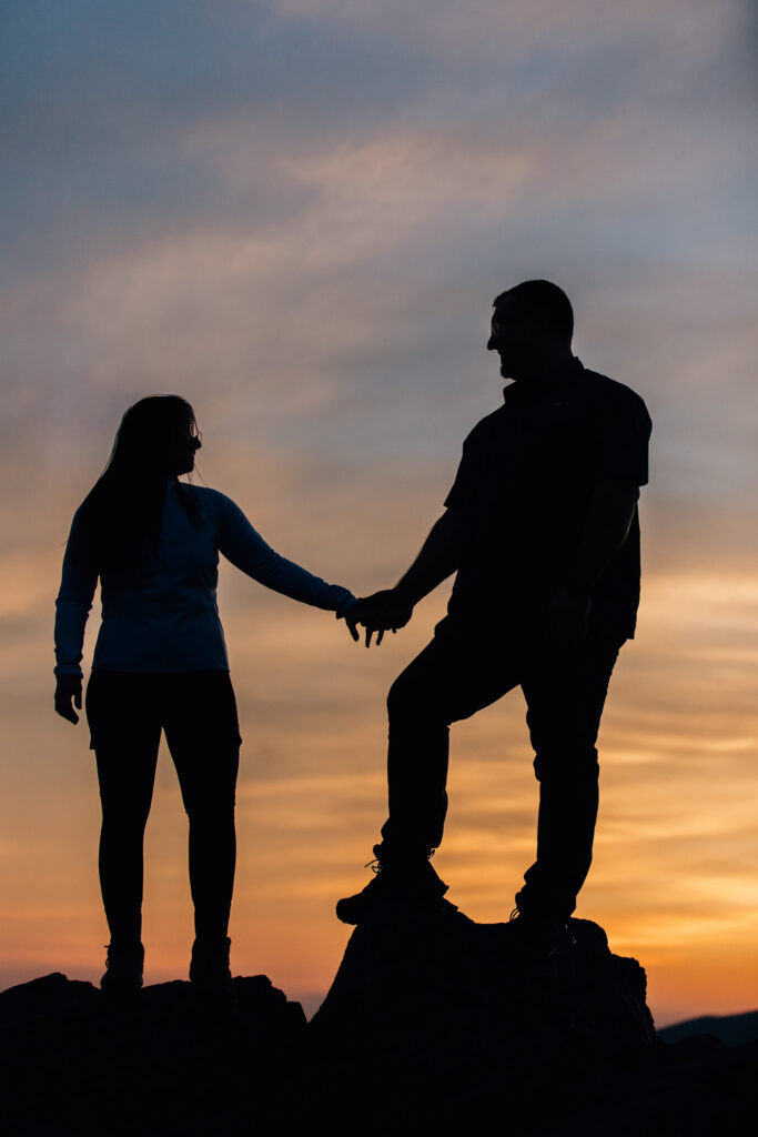 couple holds hands silhouetted against the sunset on a mountaintop after their proposal at grayson highlands state park in va