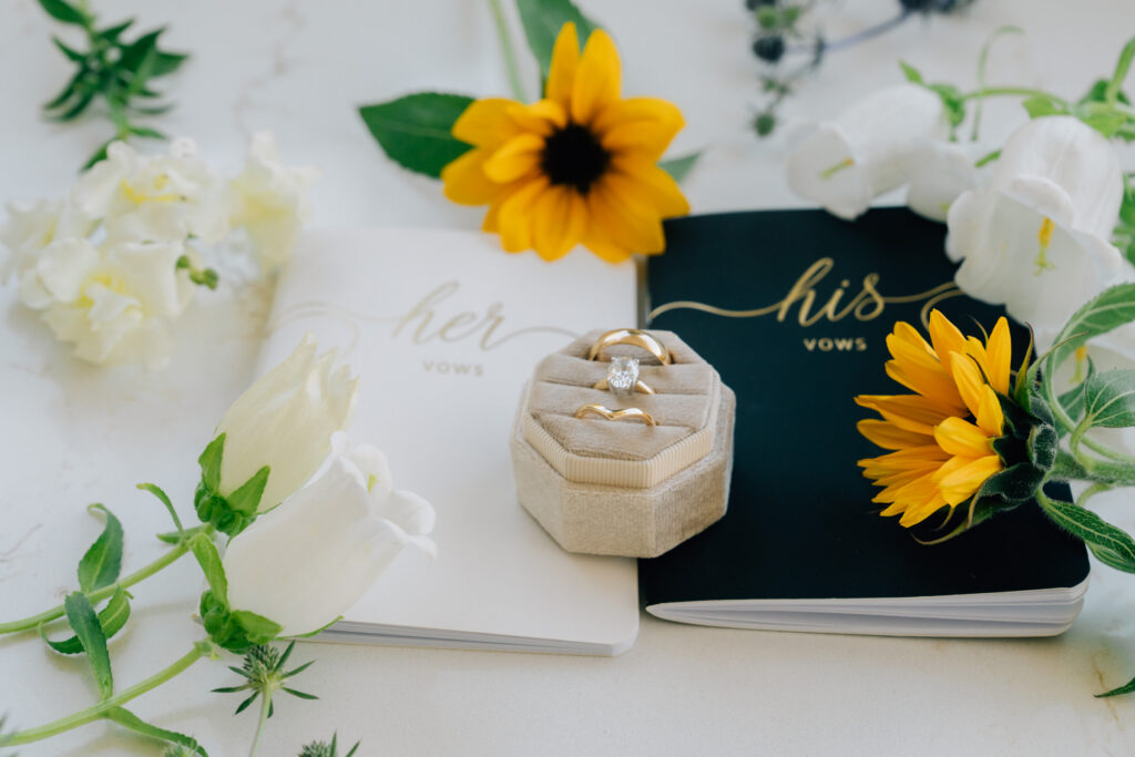 closeup photo of vow books and wedding rings surrounded by yellow and white wedding flowers