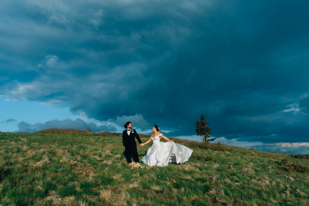 wedding couple standing on a windy mountain top at roan mountain with a dramatic stormy sky behind them while they are illuminated by the sun