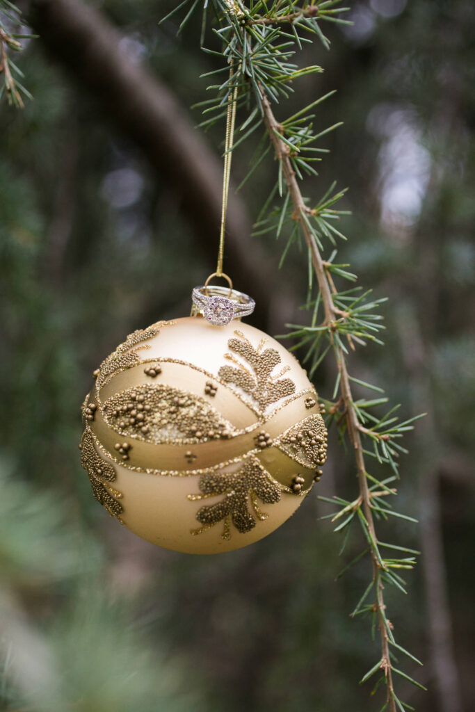 gold christmas ornament hanging from evergreen with an engagement ring on top