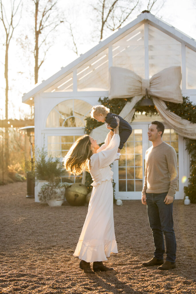 mom lifts toddler up in to the air and spins during a golden hour morning christmas family session at fleurish greenhouse in taylorsville, nc