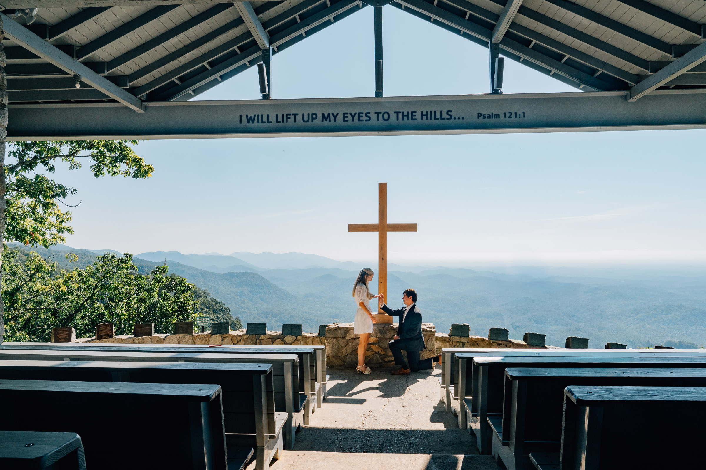 man down on one knee at symmes chapel in sc putting an engagement ring on his partner's hand in front of the big cross in the open air chapel overlooking the blue ridge mountains