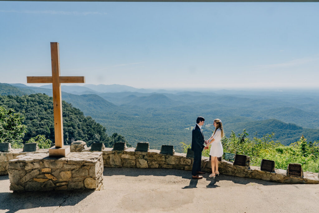 next to a large cross and with the blue ridge mountains in the background, a couple holds hands and faces one another after their proposal at pretty place chapel