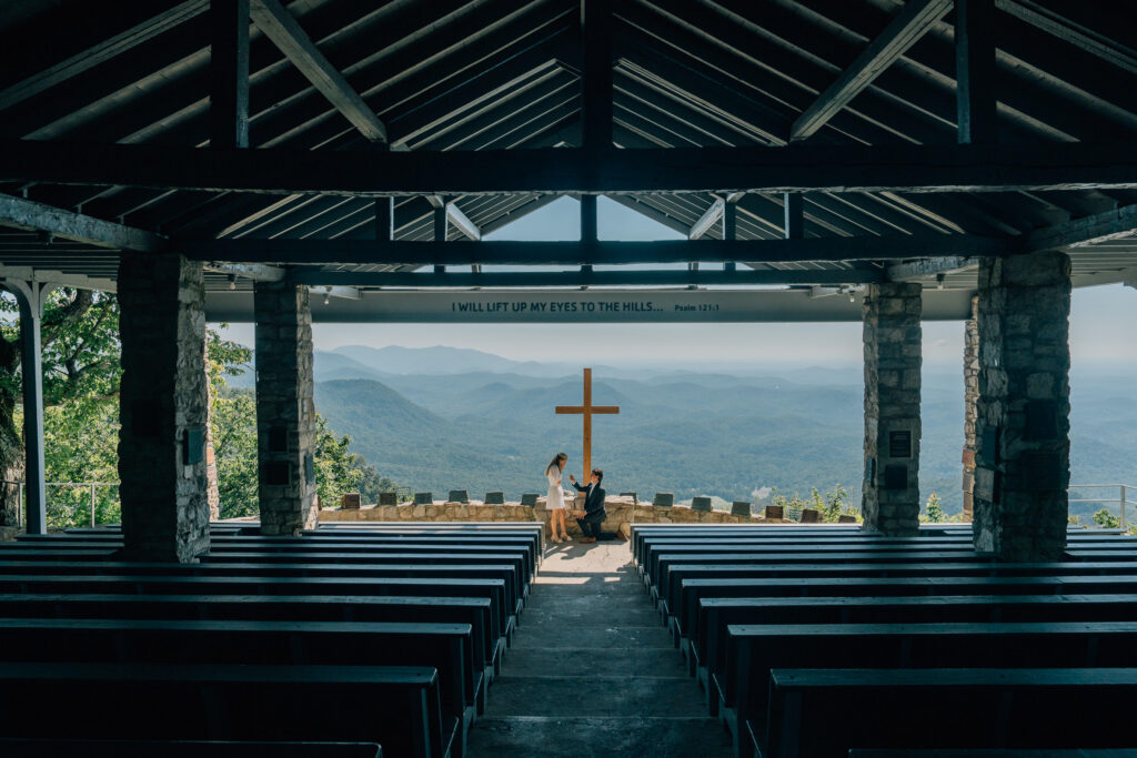 down on one knee, a man proposes to woman on one knee with an expansive outdoor chapel surrounding them and a large cross and appalachian mountains behind them