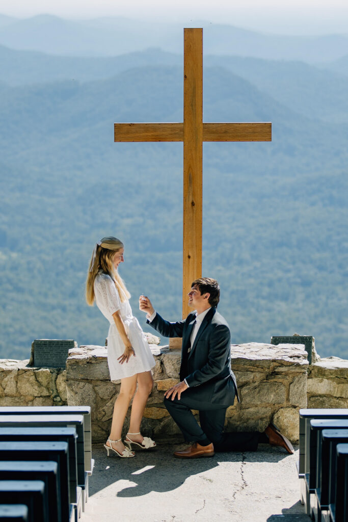 man proposes with incredible blue ridge mountain scenery behind him and a large cross at pretty place chapel while his girlfriend reacts with surprise